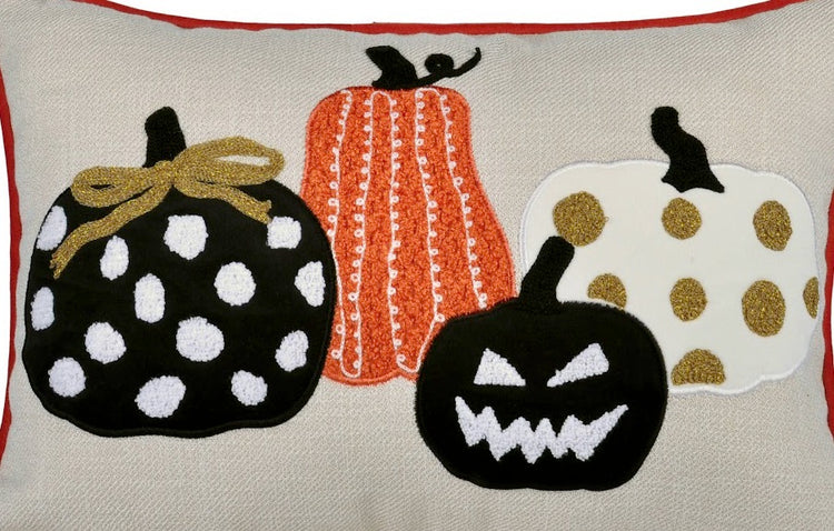 A close-up of a decorative textile or pillow featuring three stylized pumpkins: a black pumpkin with white polka dots and a gold bow, an orange pumpkin with white vertical stripes, and a white pumpkin with gold polka dots and a black stem, all on a beige background with red border trim.
