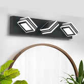 A modern three-light wall sconce with a matte black rectangular housing and white geometric, layered square-shaped bulbs, mounted on a white wall above a wooden-framed mirror with green plants in the foreground.