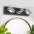 A modern three-light wall sconce with a matte black rectangular housing and white geometric, layered square-shaped bulbs, mounted on a white wall above a wooden-framed mirror with green plants in the foreground.