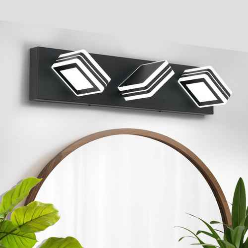 A modern three-light wall sconce with a matte black rectangular housing and white geometric, layered square-shaped bulbs, mounted on a white wall above a wooden-framed mirror with green plants in the foreground.