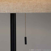 Close-up of a table lamp with a beige woven fabric shade, a black metal stem, and a black pull chain with a black weighted end, shown against a neutral gray background.