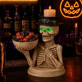 A Halloween skeleton figurine holding a bowl of candy, styled on a bar counter with a lit candle on its head and a glowing jack-o&