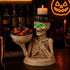 A Halloween skeleton figurine holding a bowl of candy, styled on a bar counter with a lit candle on its head and a glowing jack-o&