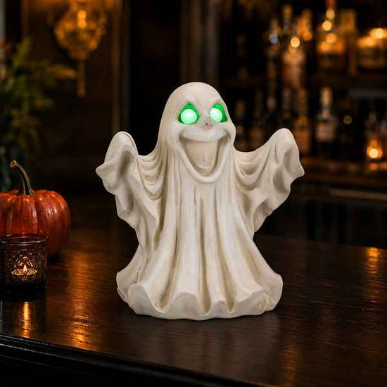 A white ghost-shaped decorative figurine with glowing green eyes sits on a dark wooden table in a dimly lit bar setting, with a small orange pumpkin and candle visible nearby.