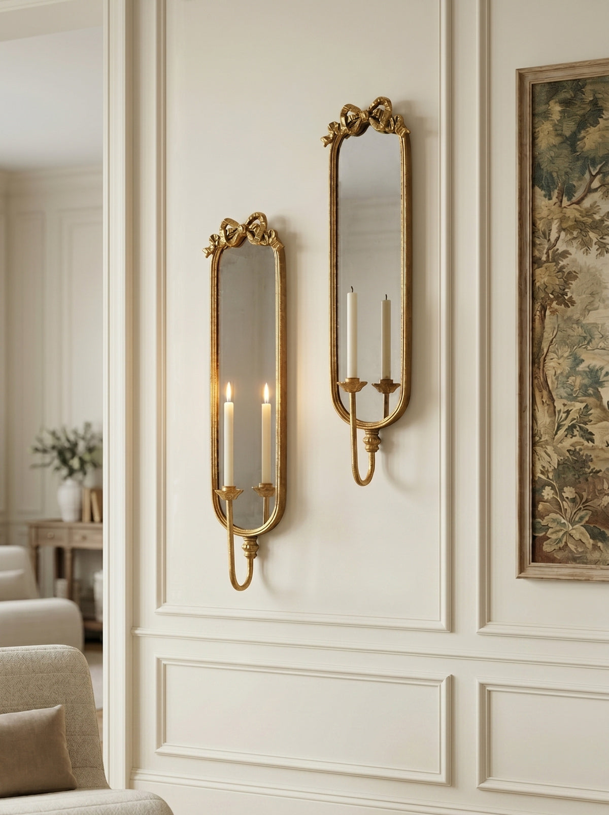 Two ornate gold wall sconces with candle-style lights mounted on a cream paneled wall next to a framed botanical print.