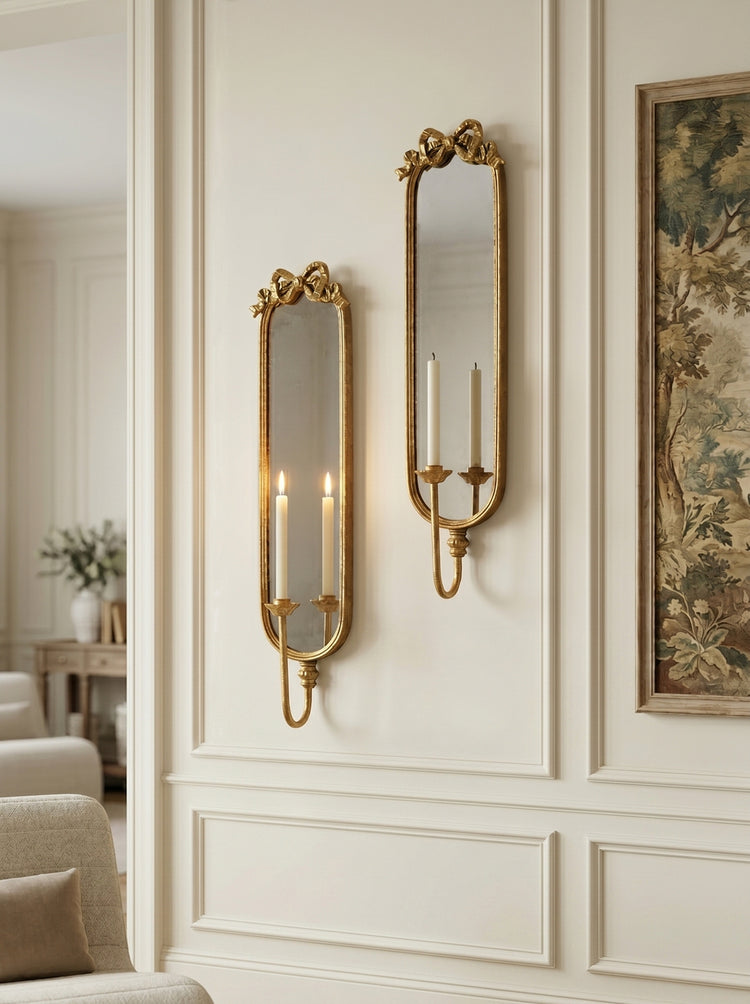 Two ornate gold wall sconces with candle-style lights mounted on a cream paneled wall next to a framed botanical print.