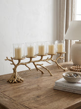 A five-light branch-shaped candle holder in a natural wood finish, styled on a rustic wooden table next to a lamp, books, and a bowl of pebbles.