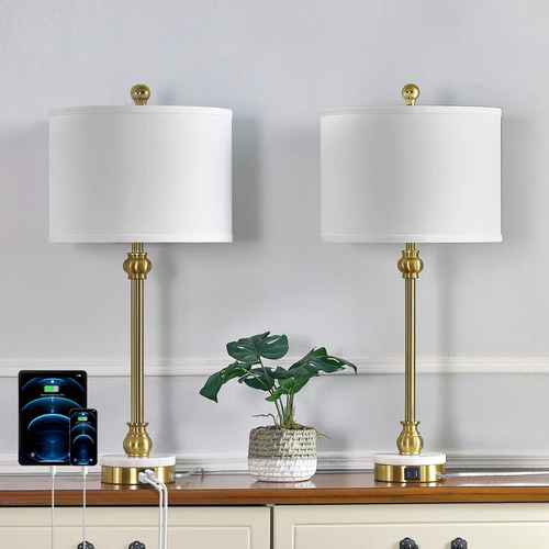 Two matching table lamps with white drum shades and gold-finished bases and stems, displayed on a wooden console against a light gray wall. A small potted green plant and a smartphone charging setup sit in front of the lamps.