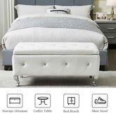 White tufted storage ottoman in front of a bed with gray bedding.