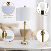 A table lamp with a white fabric drum shade, a slim gold metal stem, and a round marble-and-metal base, shown on a white table in a softly blurred interior. Four circular callouts highlight features: premium white fabric shade, £26 lamp socket, sturdy metal & marble double base, and slender column gold lamp body.