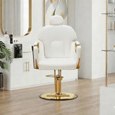 A modern white salon barber chair with gold accents and a gold hydraulic base, set in a bright, contemporary salon interior with light wood floors and white cabinets.