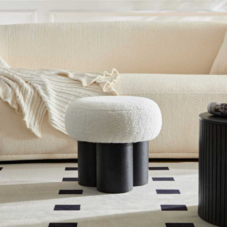 Round ottoman with black cylindrical legs in front of a beige sofa on a patterned rug.