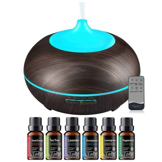 A round essential oil diffuser with a dark wood-grain finish and a bright blue illuminated top, shown with a remote control and six small essential oil bottles labeled Sweet Orange, Tea Tree, Lavender, Eucalyptus, Lemongrass, and Peppermint on a white background.
