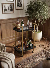 Black and gold metal bar cart with two tiers, gold frame, black trays, and casters, holding liquor bottles and glasses in a cozy home bar
