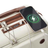 A close-up of a cream-colored hard-shell suitcase with brown leatherette trim, showing a smartphone plugged into its side charging port with a white cable. The phone screen displays a green charging icon.