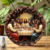 Round carved decorative wall plaque depicting the Last Supper, shown standing on a wooden surface with large green leaves in the background.