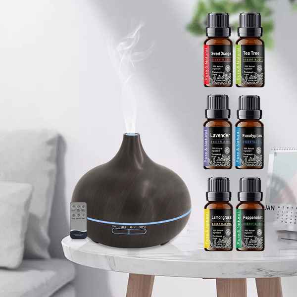 A dark brown essential oil diffuser with a remote control sits on a white marble side table in a living room setting, with steam rising from the top. On the right side of the image, six small amber glass essential oil bottles with black caps are neatly arranged in two columns, labeled with scents: Sweet Citrus, Tea Tree, Lavender, Excursion, Bergamot, and Peppermint.