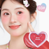 A close-up of a smiling woman with pink blush and rosy lips, holding a heart-shaped compact labeled &