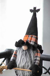 A Halloween witch gnome decoration with a black felt hat topped with a bat silhouette, an orange, black, and white plaid band, a long gray faux-fur beard, and black gloves, holding a broom.