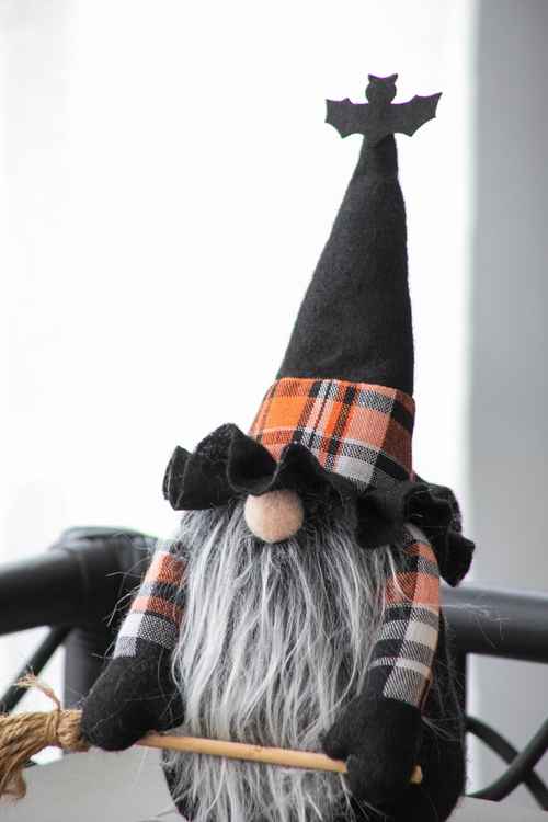 A Halloween witch gnome decoration with a black felt hat topped with a bat silhouette, an orange, black, and white plaid band, a long gray faux-fur beard, and black gloves, holding a broom.