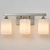 A three-light wall sconce with a brushed metal bar and cylindrical frosted glass shades, shown illuminated against a plain light wall.