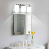A three-light bathroom vanity set with a rectangular mirror and a wall-mounted chrome light fixture above a white vanity cabinet. The lights are turned on, casting a soft glow, and the mirror reflects a bathroom interior.