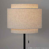 A modern floor lamp with a black metal stand and three horizontal drum shades made of textured beige linen, shown against a neutral gray background. A pull chain is visible at the bottom.