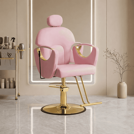 A pink salon barber chair with gold metal accents and a circular gold base, shown in a modern spa-like interior with a mirror, shelving, and a vase.