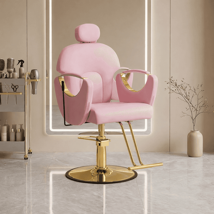 A pink salon barber chair with gold metal accents and a circular gold base, shown in a modern spa-like interior with a mirror, shelving, and a vase.