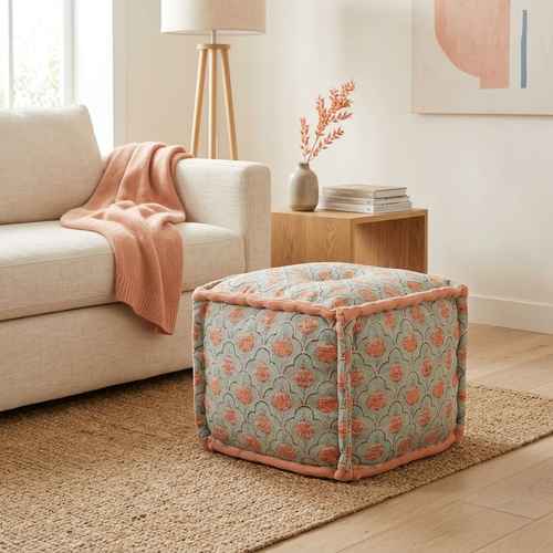A square upholstered ottoman with a muted sage green floral pattern and peach-pink piping, styled in a bright living room next to a beige sofa.