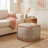 A square upholstered ottoman with a muted sage green floral pattern and peach-pink piping, styled in a bright living room next to a beige sofa.
