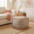 A square upholstered ottoman with a muted sage green floral pattern and peach-pink piping, styled in a bright living room next to a beige sofa.