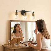 A woman applies makeup while looking into a wooden vanity mirror, with an orange tabby cat on the dresser below. A black three-light wall sconce with brass-finished sockets is mounted above the mirror.