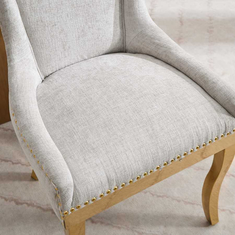 Close-up of a gray upholstered chair with wooden legs and gold nailhead trim on a light background.