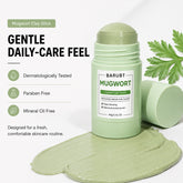 BARUBT Mugwort Clay Stick shown in a light green container with the cap off, placed on top of a smear of pale green clay product. A second cap lies beside the container, and a green mugwort leaf is visible at the upper right.