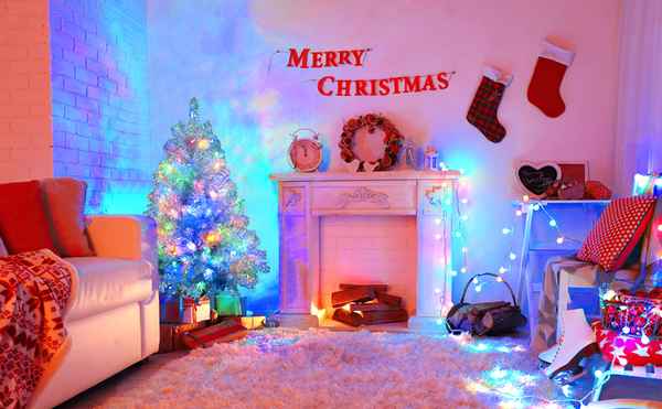 A cozy Christmas living room decorated with a lit Christmas tree, fireplace mantel with stockings and wreath, colorful string lights, and wrapped gifts on a white shag rug.