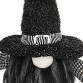A close-up of a black witch-themed decorative figure or doll with a tall glittery black hat, a black-and-white striped band with a silver buckle, black hair, and a white face on a white background.