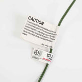 A close-up of a green electrical wire with two tags attached on a plain white background. The top tag is a white caution label with black text, and the bottom tag is a white UL certification label with red and black text.