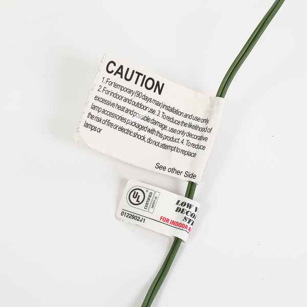 A close-up of a green electrical wire with two tags attached on a plain white background. The top tag is a white caution label with black text, and the bottom tag is a white UL certification label with red and black text.