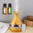 A wooden-patterned essential oil diffuser with a purple band and a white remote control, shown on a tabletop with two small fragrance bottles and a gray wall behind it.