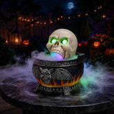 Halloween decoration set featuring a skull with glowing green eyes and a multicolor lighted mouth, emerging from a black cauldron with bat and flame details, placed on a round table with fog effects in a decorated backyard setting.