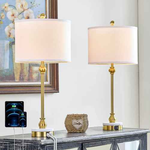 Two matching table lamps with white drum shades and gold-finished metal bases are shown on a console table in a styled interior. One lamp has a white circular base, and the other has a white base with a small gold accent.