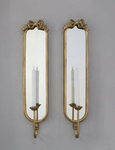 A pair of ornate gold wall sconces with elongated rectangular mirrors and candle-style light holders, mounted against a plain gray wall.
