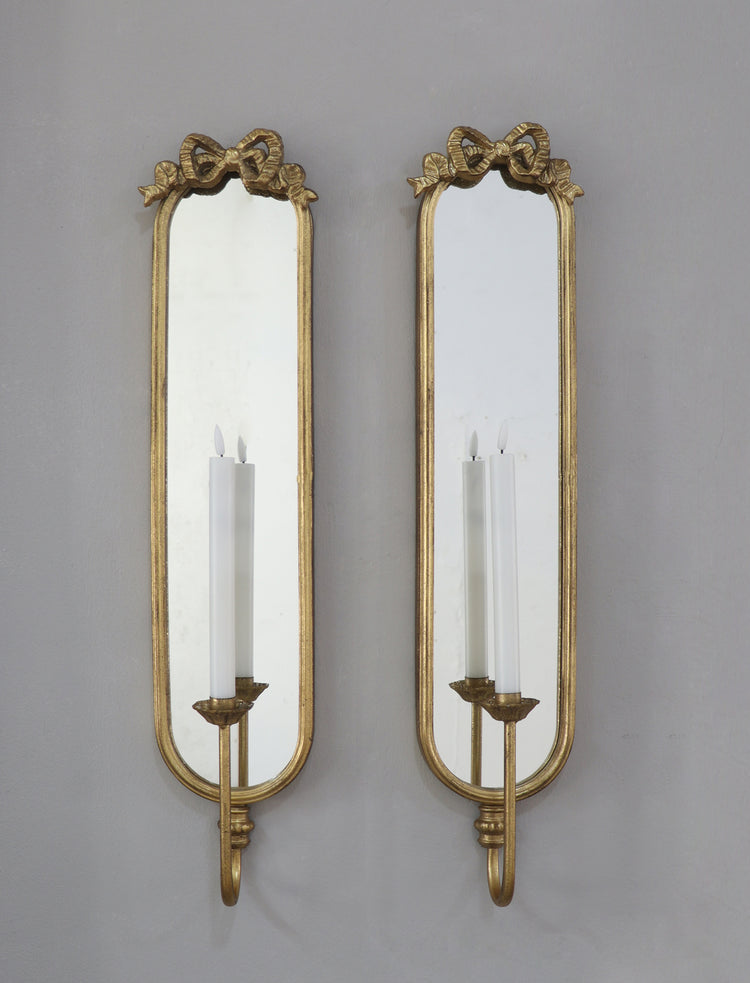 A pair of ornate gold wall sconces with elongated rectangular mirrors and candle-style light holders, mounted against a plain gray wall.