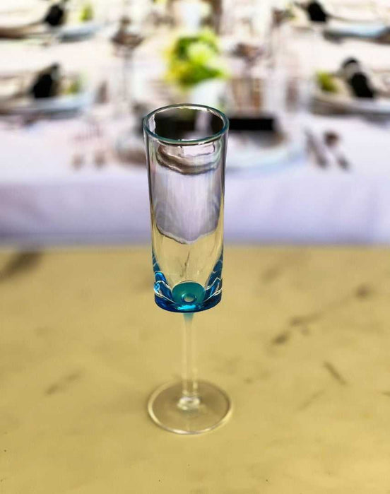 Single blue acrylic unbreakable champagne flute from the set of 4, shown on a table with a formal dining setup in the background.