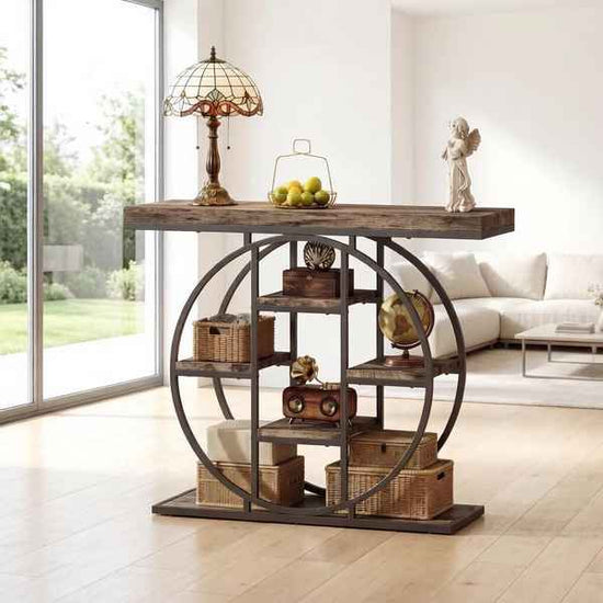 A rustic dark wood and metal console table styled in a bright, modern living room. The table features a thick distressed dark wood top, a large circular metal frame, and multiple open shelves holding wicker baskets, a vintage radio, a globe, and decorative items.