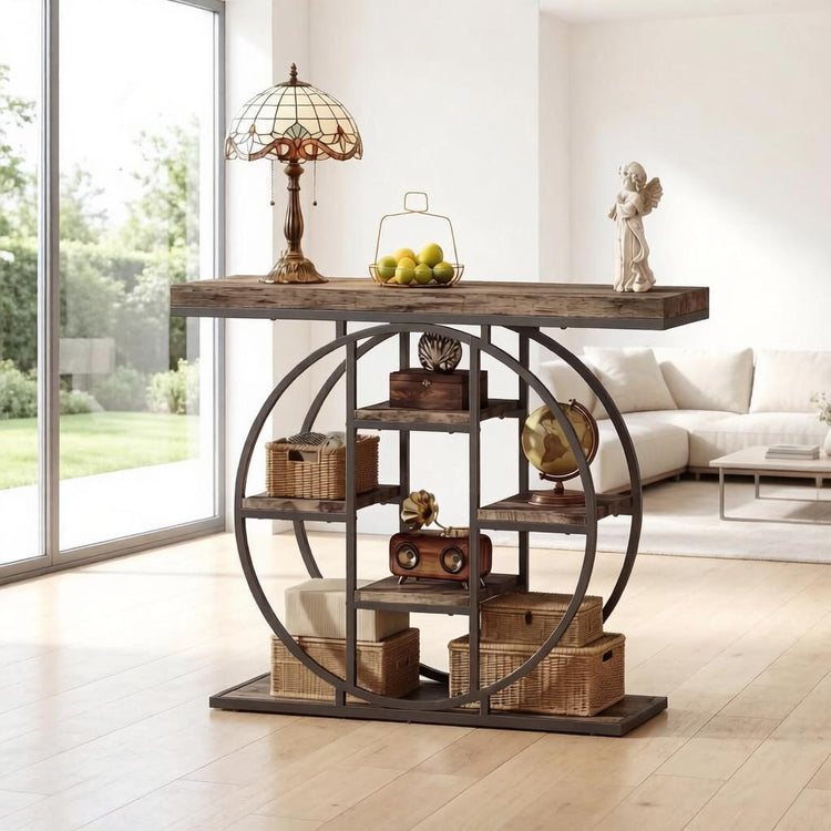 A rustic dark wood and metal console table styled in a bright, modern living room. The table features a thick distressed dark wood top, a large circular metal frame, and multiple open shelves holding wicker baskets, a vintage radio, a globe, and decorative items.