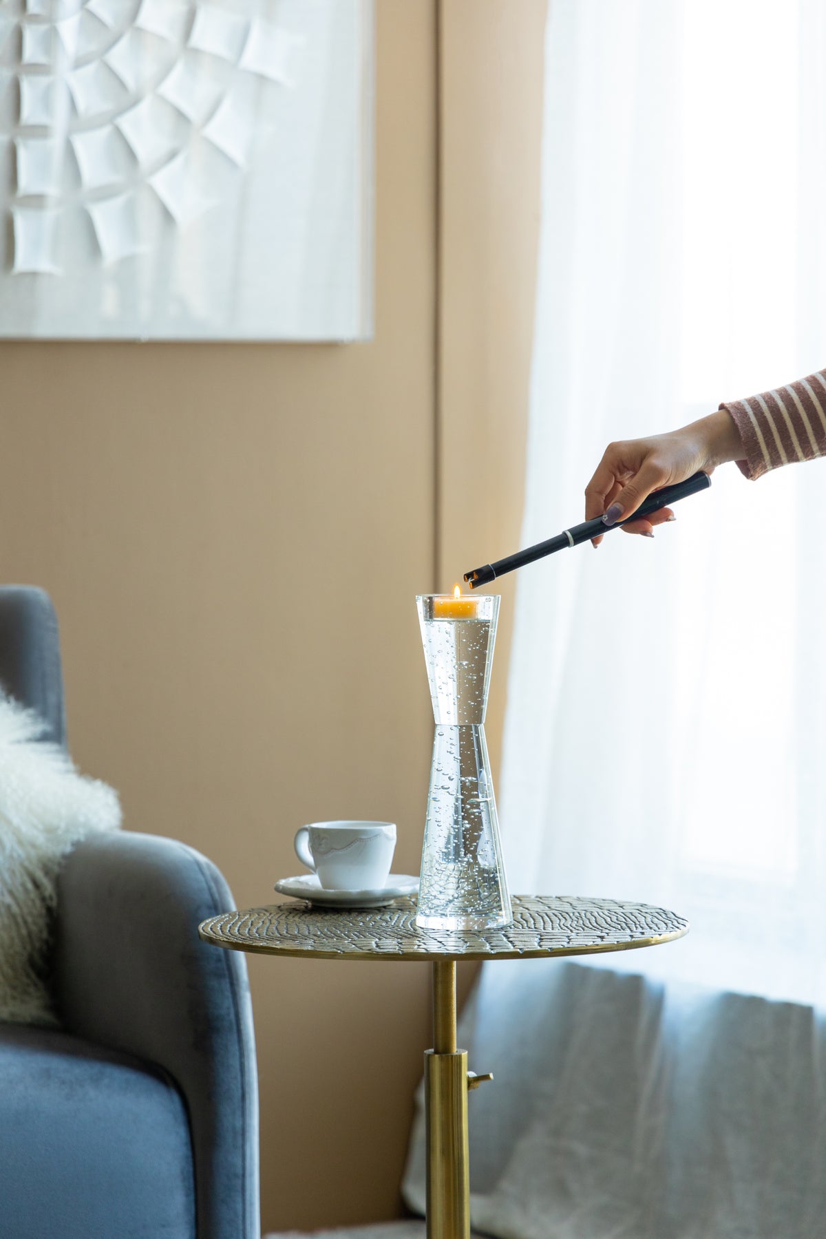 A hand holding a black lighter ignites a small orange candle inside a clear, textured glass vase set on a round woven side table in a modern living room.