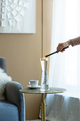 A hand holding a black lighter ignites a small orange candle inside a clear, textured glass vase set on a round woven side table in a modern living room.
