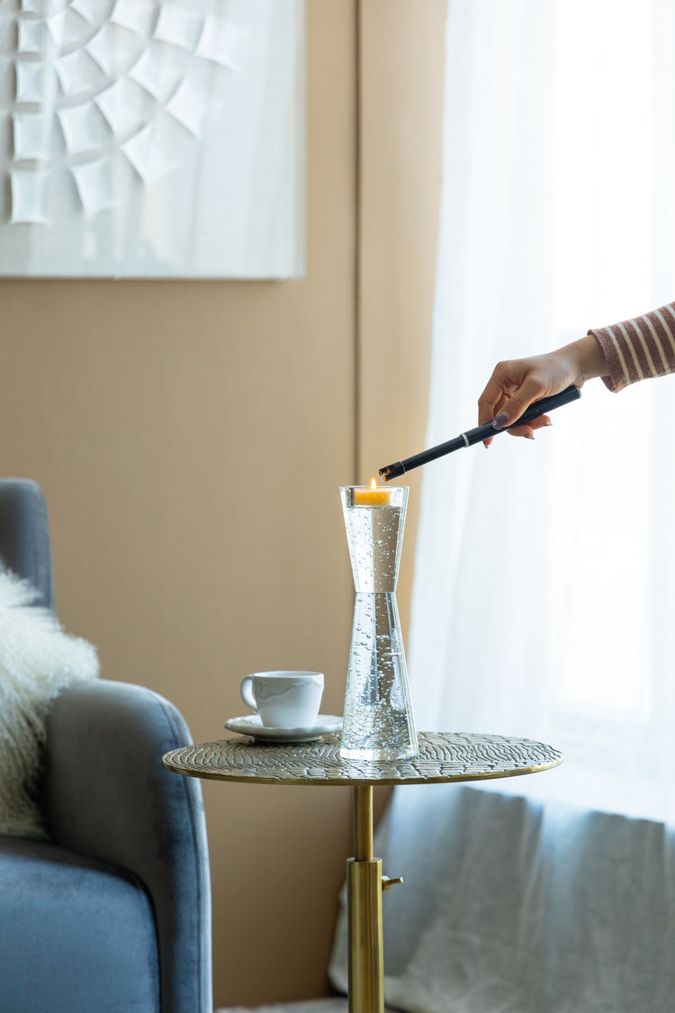 A hand holding a black lighter ignites a small orange candle inside a clear, textured glass vase set on a round woven side table in a modern living room.